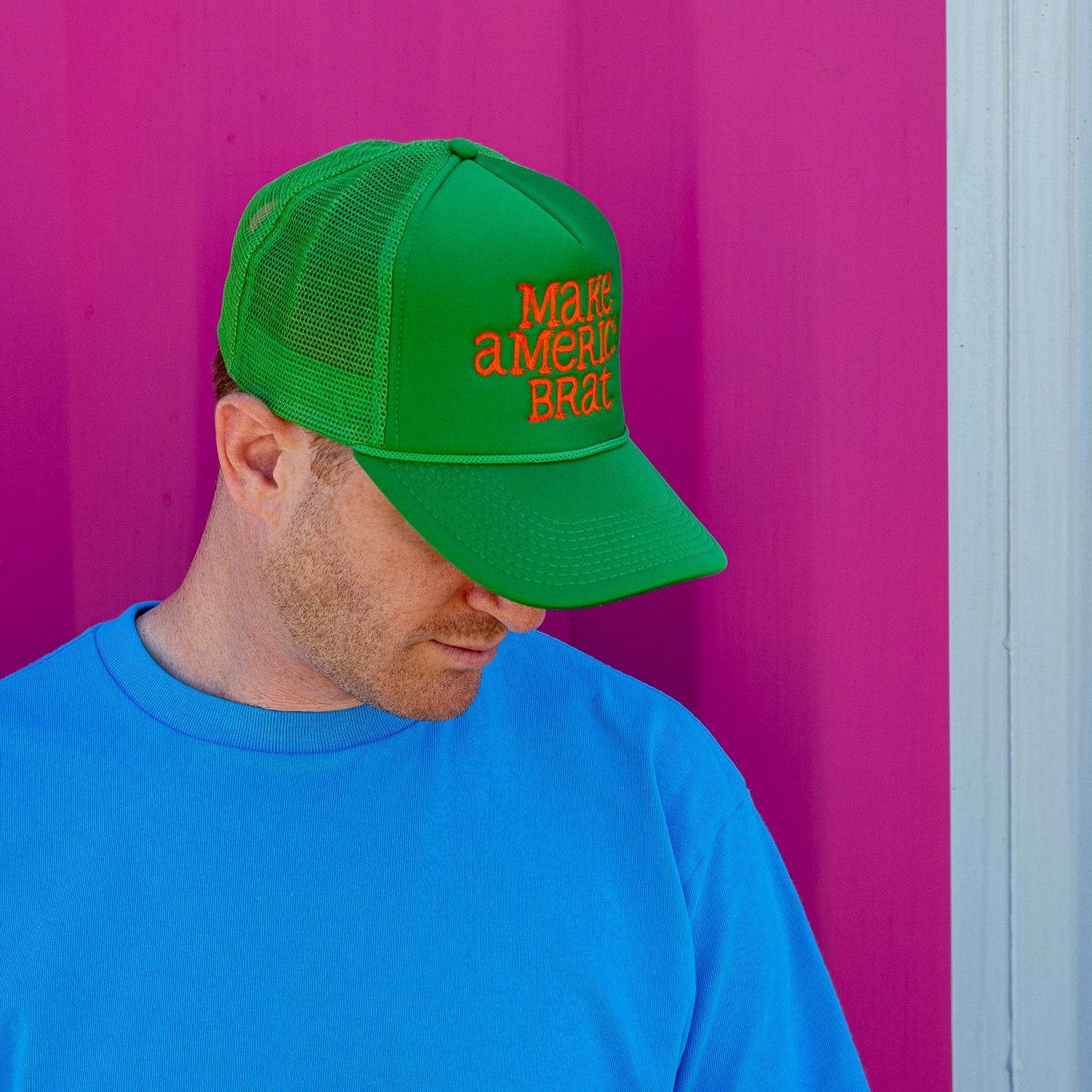 Man standing in front of pink shipping container wearing green and orange Make America Brat Hat.