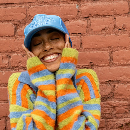 Woman standing in front of red brick wall, smiling and holding her face wearing Make America Dope Sky Blue Hat.