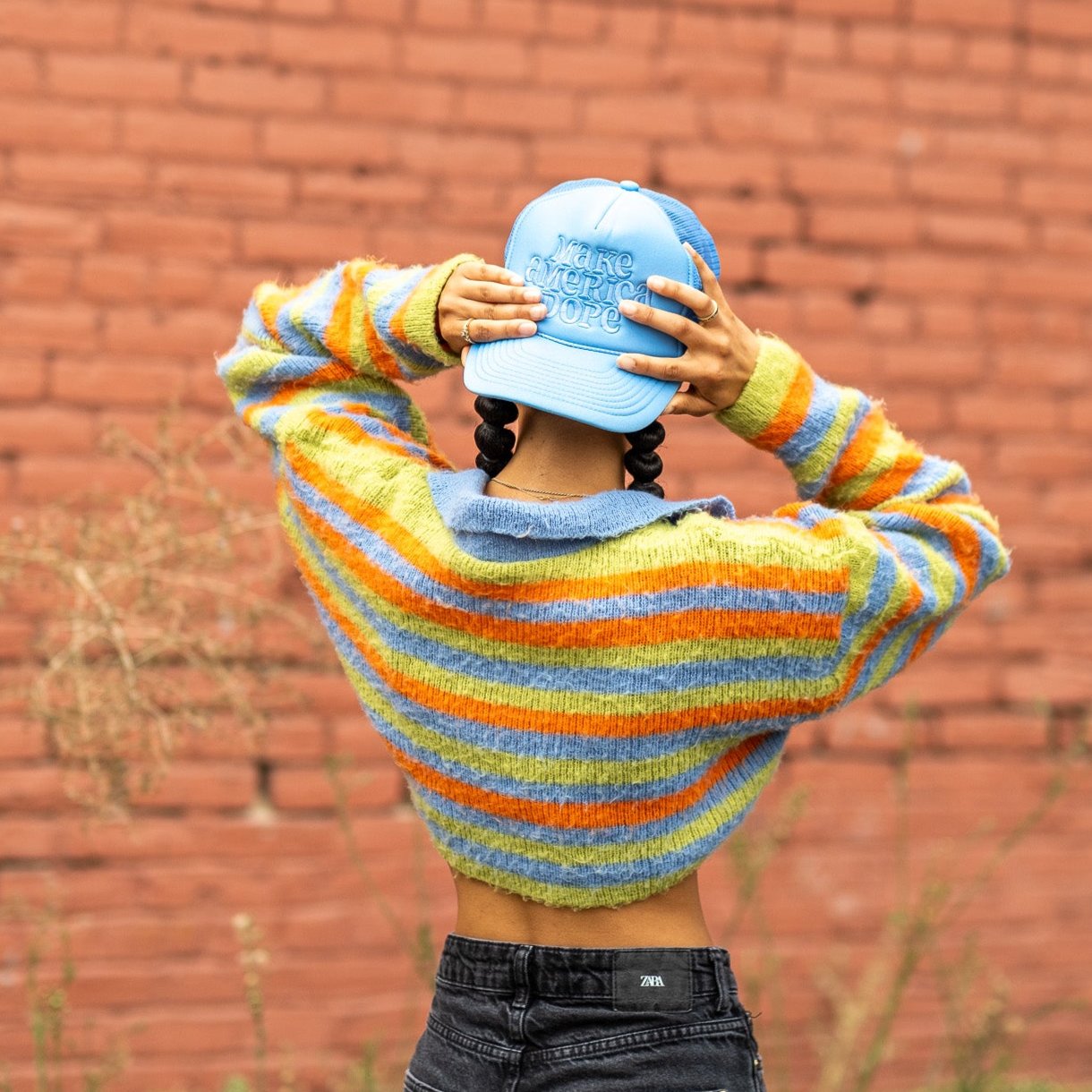 Woman standing in front of red brick wall wearing colorful sweater and Make America Dope Sky Blue Hat